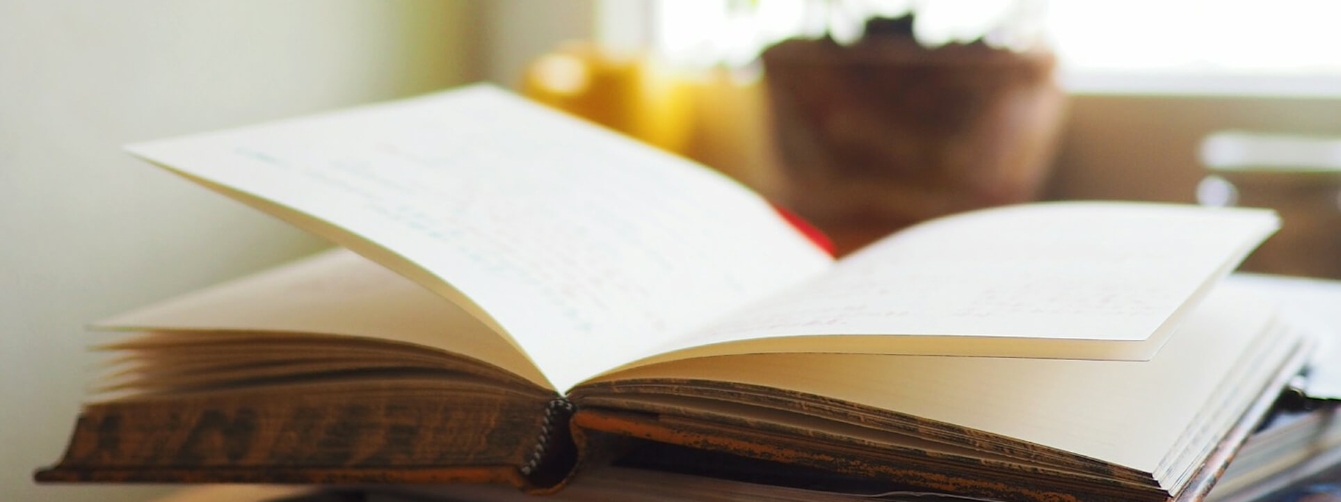 木製の机の上に開かれた厚みのある本を、やわらかな逆光が照らしている様子。ページがめくられた瞬間の静かな読書シーンを表現した画像。