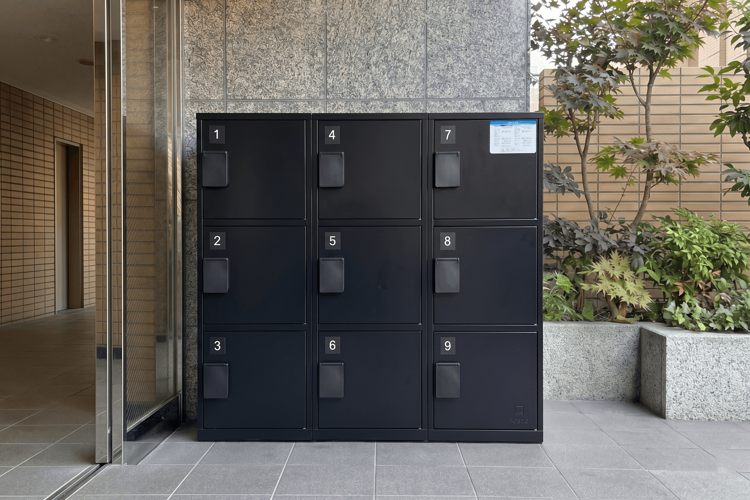 マンションの共用部に設置された黒色の宅配ボックスで、番号付きの9つの扉が整然と並ぶ収納設備